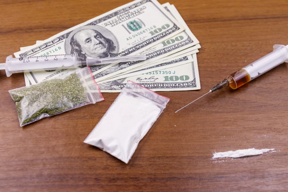 Variety of drugs including cocaine, heroin syringe, dried cannabis, and dollars on a table, representing drug use, trafficking, crime, and addiction concept.