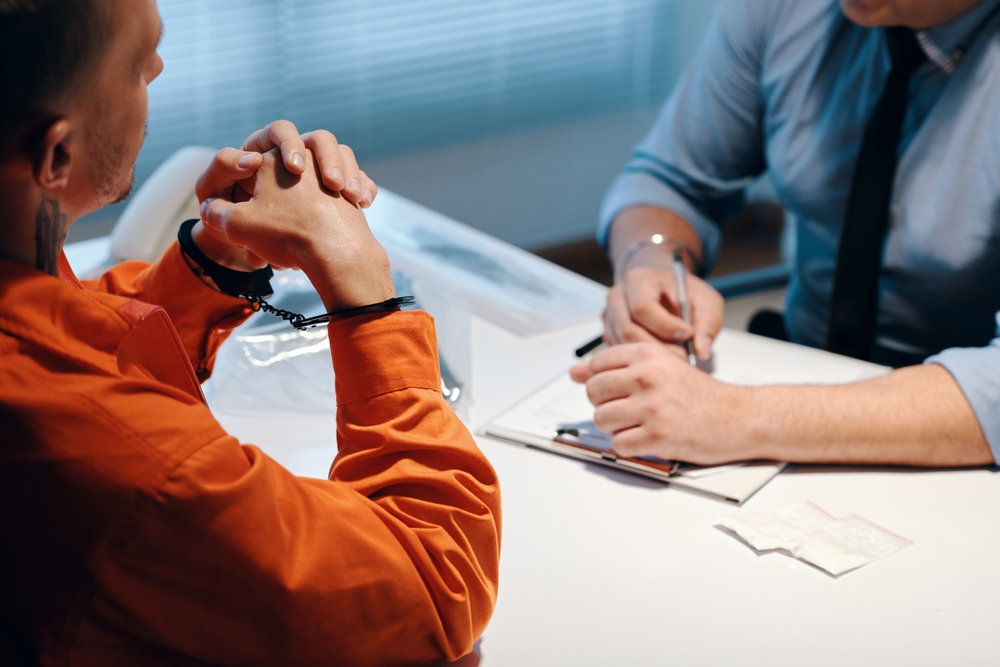 A criminal in handcuffs converses with a detective who is filling out witness statement forms.