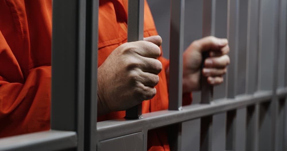 Close-up of an elderly prisoner's hands gripping metal bars, dressed in an orange uniform, standing in a prison cell. The inmate serves a sentence for a crime, confined behind bars.