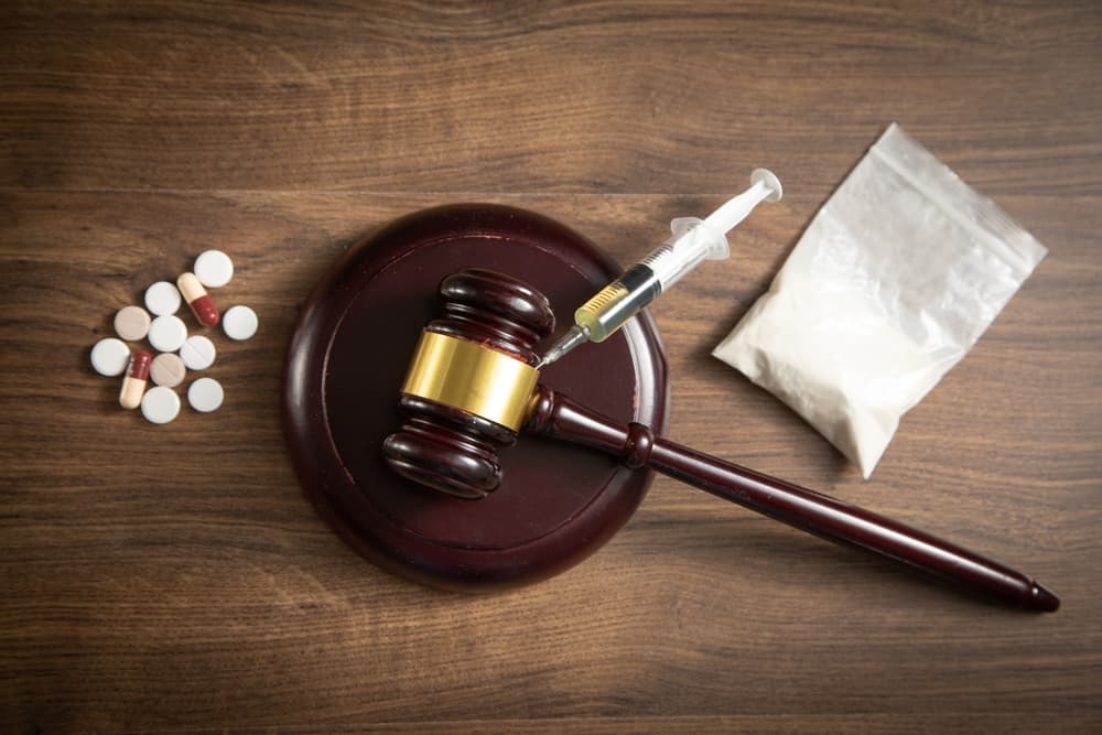 A judge's gavel is placed beside a syringe, a packet of heroin, and various drugs, symbolizing the intersection of law and the ongoing battle against drug-related crimes.