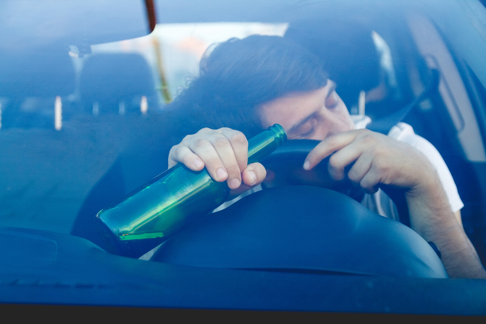 A young boy driving a car while holding a bottle of beer. The image emphasizes the dangers of drinking and driving, illustrating the concepts of DUI and DWI.






