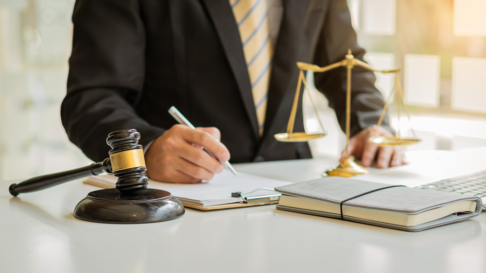 A male attorney or judge advisor in a meeting with clients, discussing legal matters. The scene includes a gavel and scales of justice on the desk, representing fairness and legal guidance.