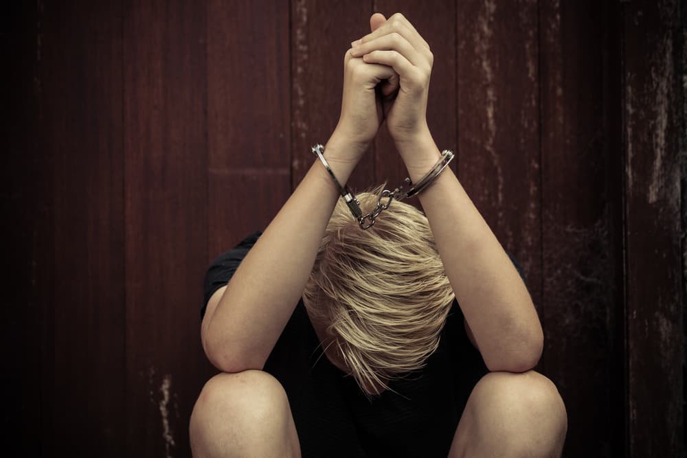 Young teenage boy wearing handcuffs sitting with his head bowed between his legs