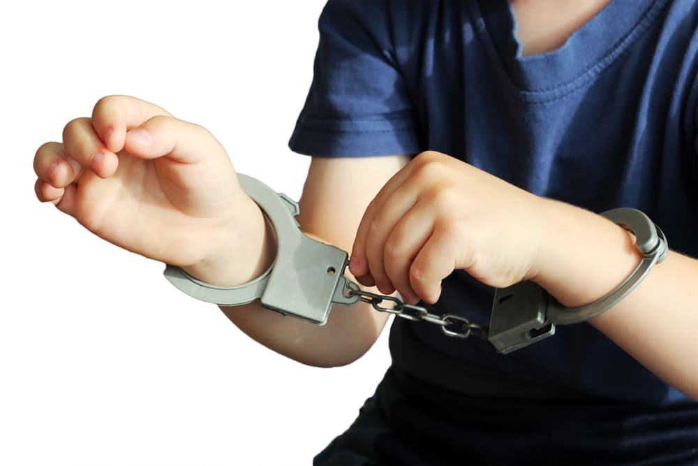 Children's hands in handcuffs, with a small toy placed nearby, isolated against a plain background.