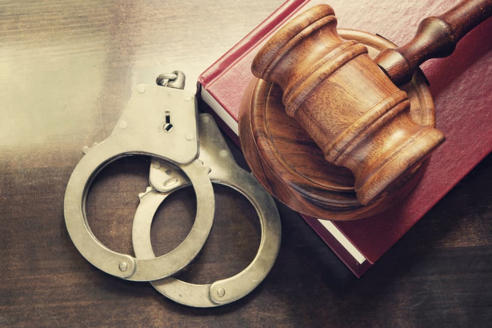 A gavel and handcuffs placed atop a red legal book on a wooden table.






