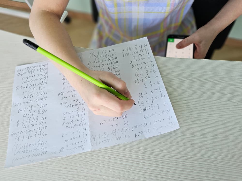 A student child discreetly holds a smartphone under the desk during an exam, attempting to cheat by using a cheat sheet or searching for answers online.